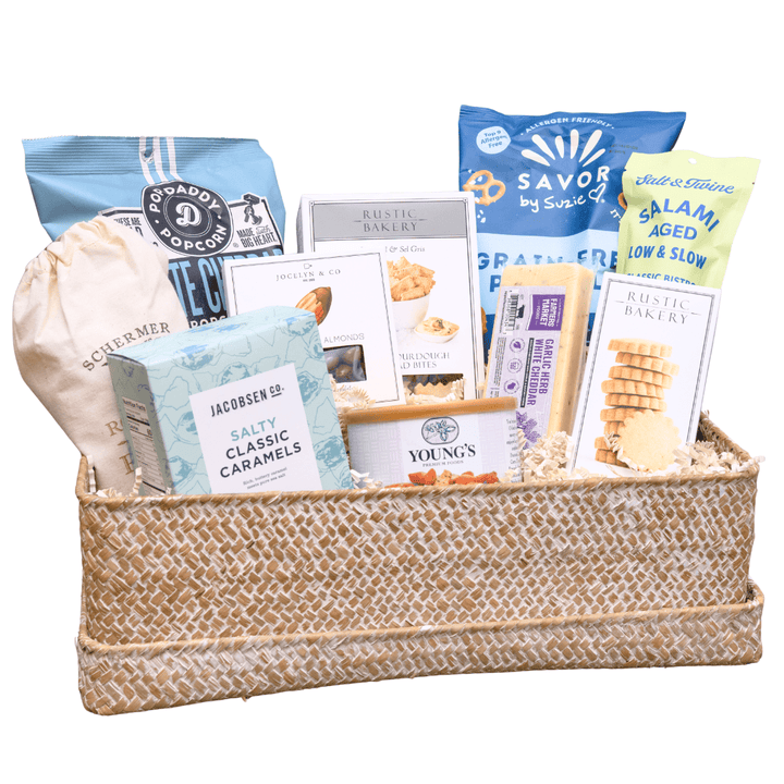 A woven natural-fiber gift basket filled with assorted packaged snacks and baked goods, including bags of chips, crackers, caramels, and pastries, photographed on a plain white background.