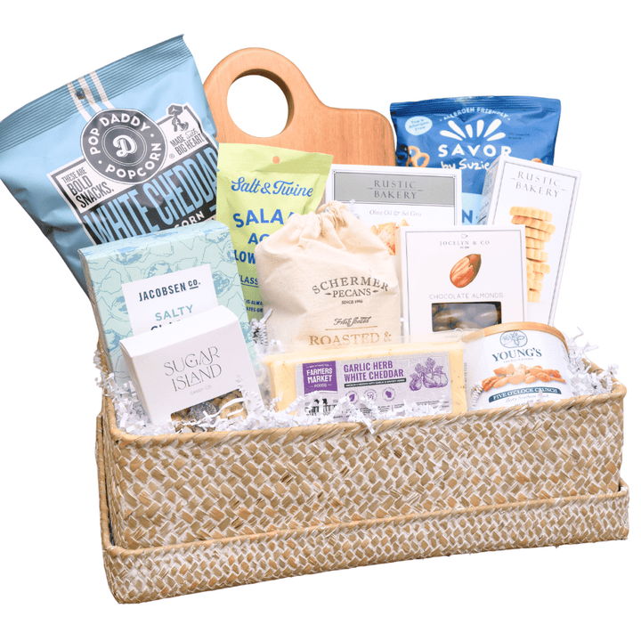 A gourmet gift basket filled with assorted snacks, cheeses, and packaged foods, set in a woven straw basket with a wooden handle on a white background.