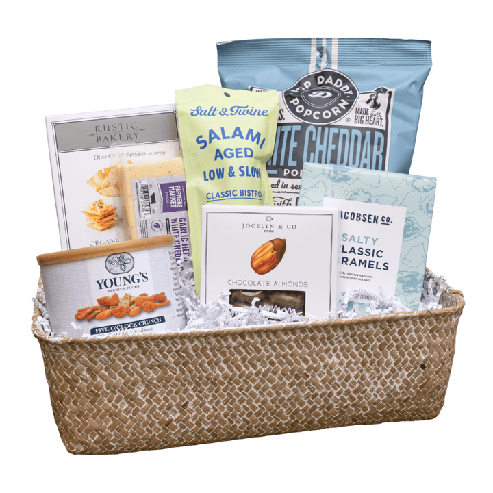 A gourmet gift basket featuring an assortment of snacks, cheeses, and baked goods arranged inside a woven natural fiber basket with white crinkle paper filler. Visible items include a bag of Poppyseed chips, a package of aged salmon snack chips, a box of classic cheddar cheese, a tin of Young's Almonds, and a bag of classic camels.