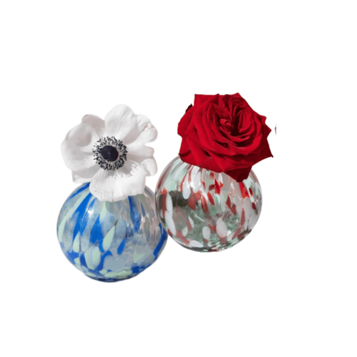 Two decorative glass vases on a plain white background, one with a blue and white marbled body and a white flower with a dark center, and the other with a red, white, and green marbled body and a red rose.