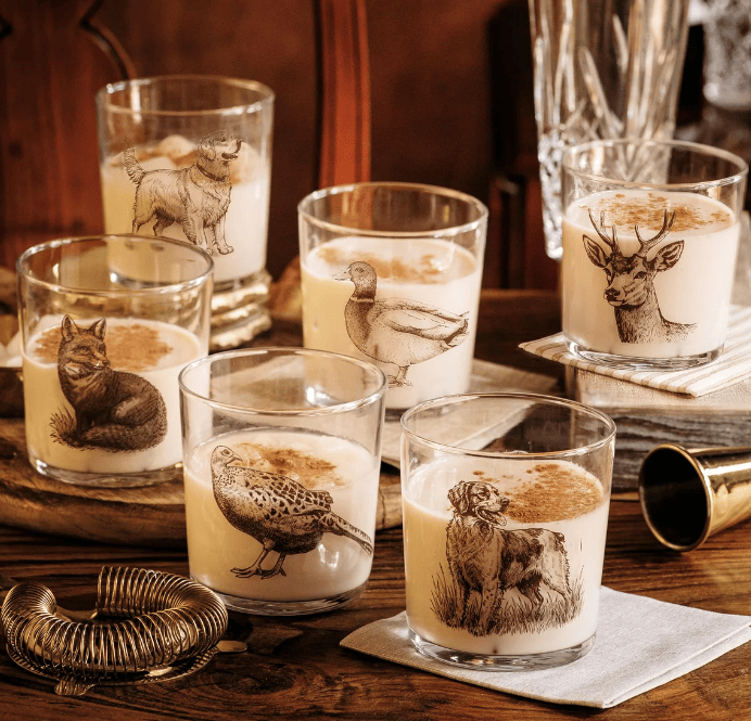 
Six wildlife themed rocks glasses filled with a creamy beverage and decorated with sepia animal illustrations, arranged on a wooden table with glasses and serving props.