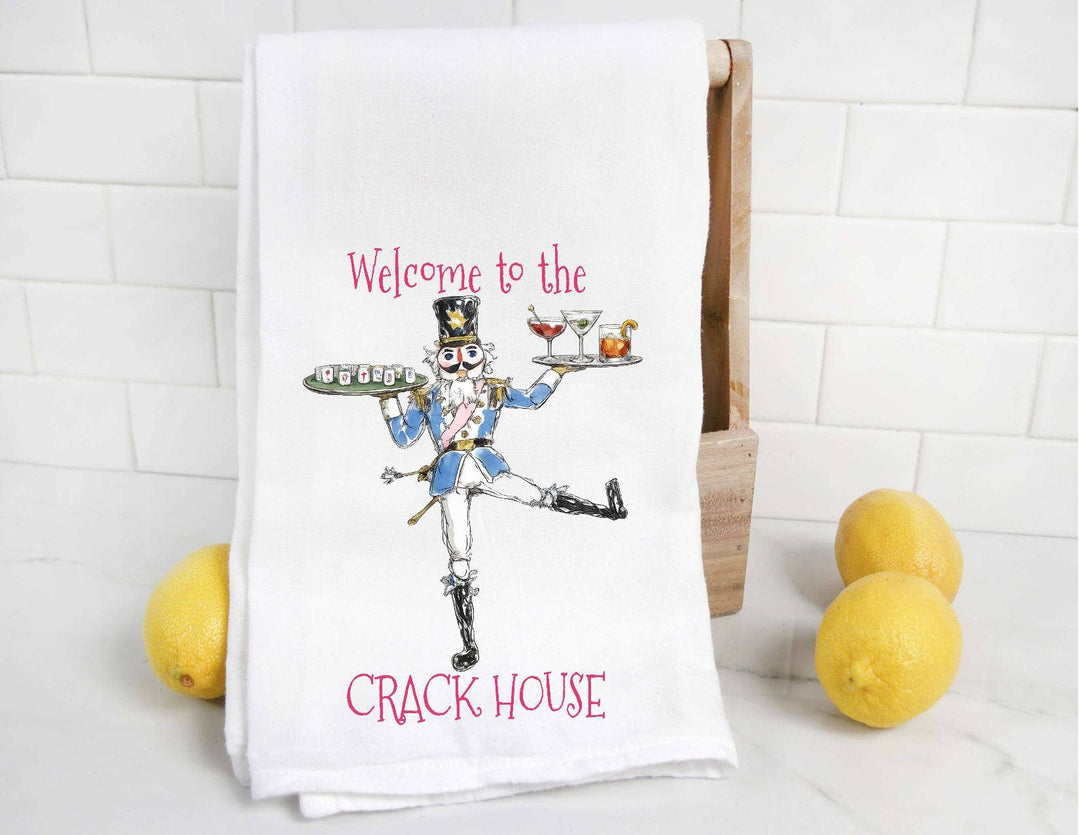 A white kitchen towel with a colorful illustration of a nutcracker waiter and the text 'Welcome to the CRACK HOUSE' in pink, displayed on a wooden stand with lemons against a white subway tile background.