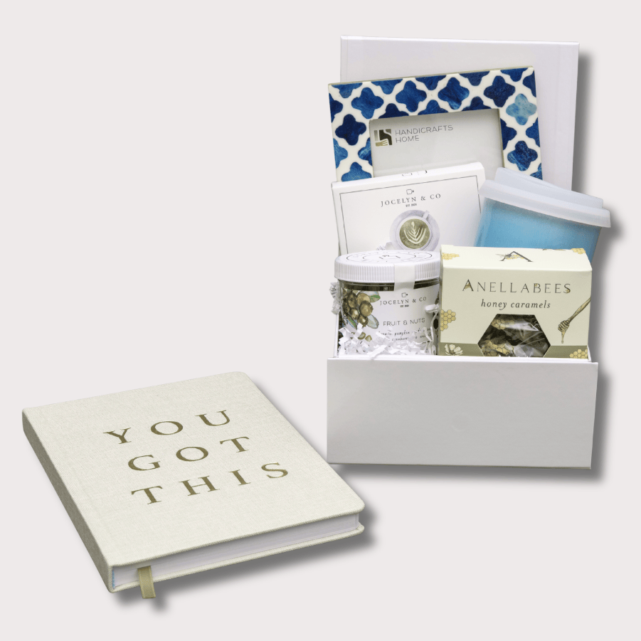 A curated gift set arranged on a light gray background, featuring a cream hardcover book with the words 'YOU GOT THIS' in gold lettering, a white gift box containing a blue-and-white patterned item, a small jar, a candle, and a boxed product labeled 'ANELLABEES'.