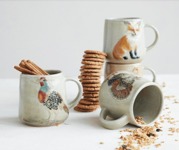 Three handmade ceramic mugs with painted animal designs, styled with cookies, cinnamon sticks, and cereal on a white surface against a light gray background.