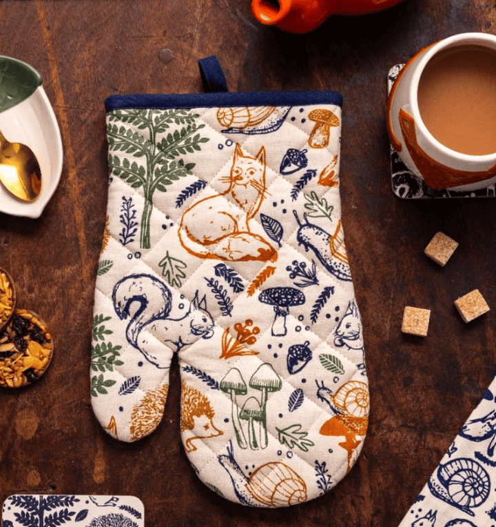 A quilted oven mitt with a woodland animal and mushroom pattern, displayed on a rustic dark wooden table alongside a cup of coffee, sugar cubes, and a small dish.