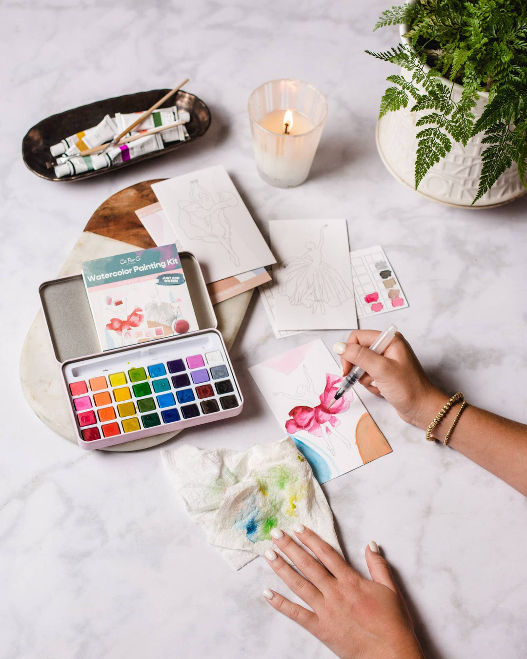 Person painting with watercolors on a table with art supplies and a candle.