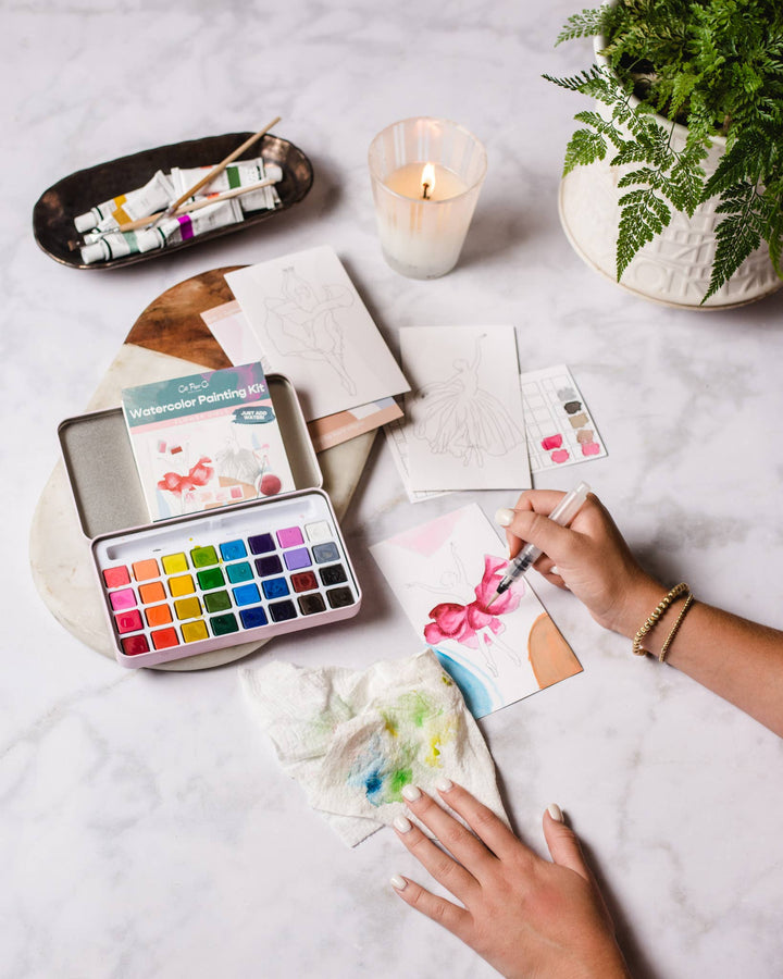 Person painting with watercolors on a table with art supplies and a candle.