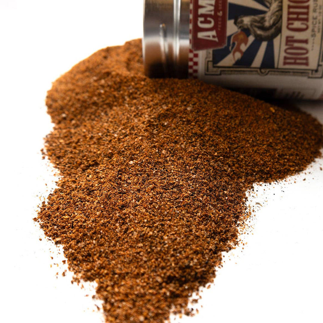 A close-up of a pile of coarse brown seasoning or spice powder spilled from a silver and white container on a white background. The container label shows partial text including 'ACM' and 'HOT CHICKEN'.
