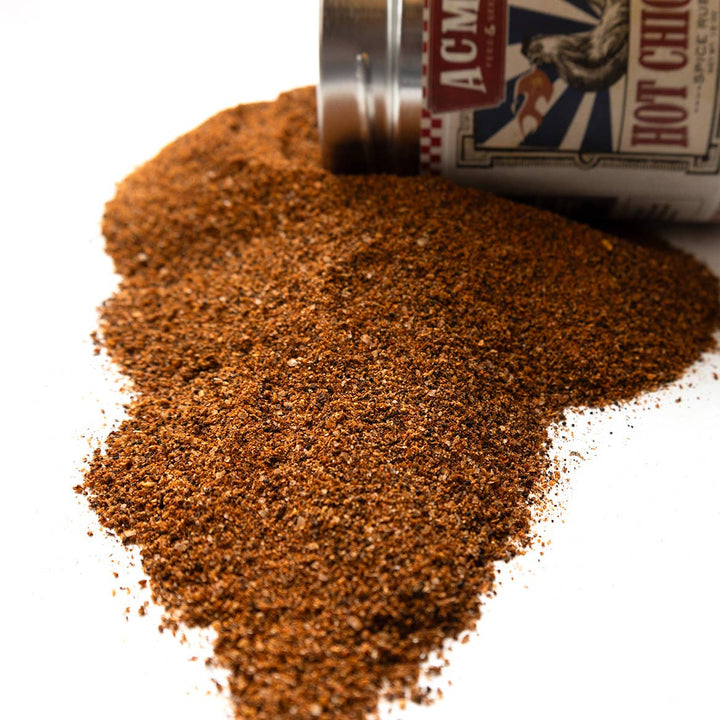 A close-up of a pile of coarse brown seasoning or spice powder spilled from a silver and white container on a white background. The container label shows partial text including 'ACM' and 'HOT CHICKEN'.