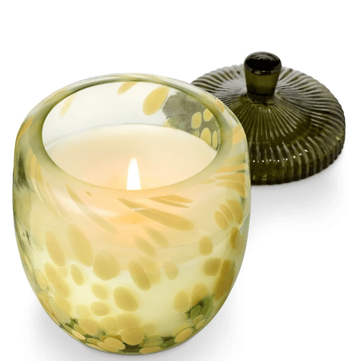 A lit glass candle in a rounded, translucent pale yellow container with green and yellow spotted leaf-like patterns, shown with its acorn lid on a white background.