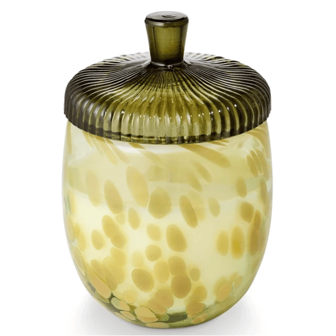 A glass candle in a rounded, translucent pale yellow container with green and yellow spotted leaf-like patterns, shown with its acorn lid on a white background.
