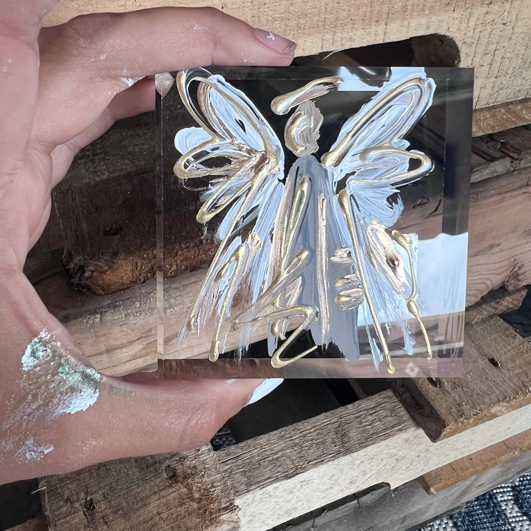 Hand holding a clear acrylic block with a gold and silver angel design on a wooden surface.