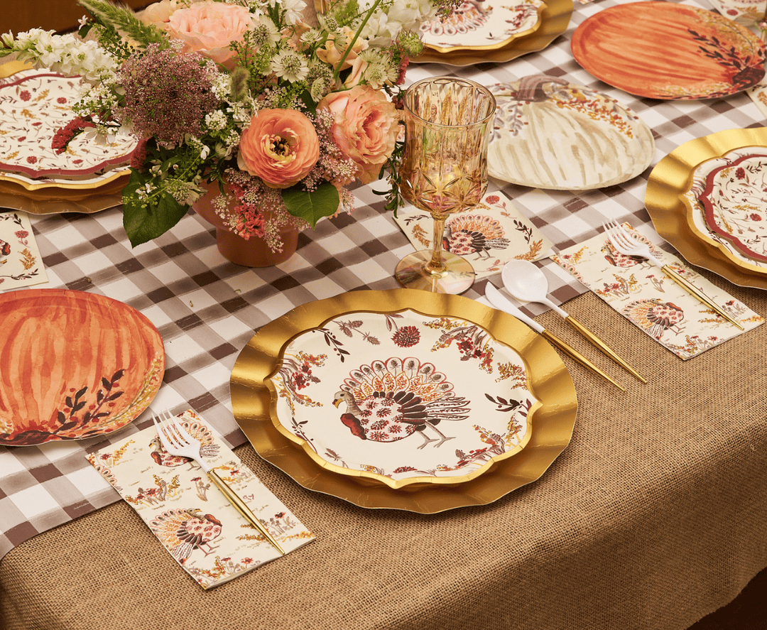 A styled Thanksgiving table setting featuring cream plates with a detailed turkey and floral pattern, layered with gold-rimmed plates, matching patterned napkins, gold flatware, and a rustic burlap tablecloth with a checkered runner and floral centerpiece.