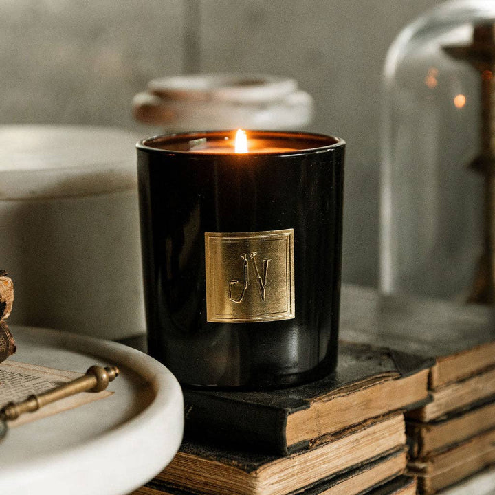 Black candle with a gold logo on a stack of books, surrounded by decorative items.
