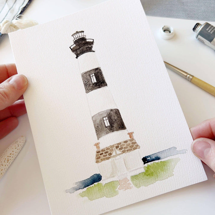 Hand holding a watercolor painting of a lighthouse on a white surface with art supplies.