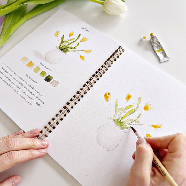Person drawing flowers in a notebook with a paintbrush, surrounded by art supplies on a white surface.