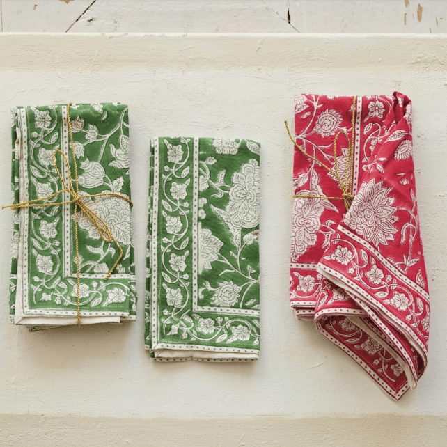 Three folded patterned cloth napkins tied with twine, displayed on a textured cream surface. Two napkins are green with white floral motifs, and one is red with white floral motifs.