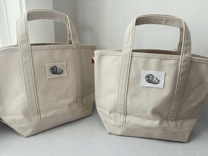 Two canvas tote bags in a light beige color, standing side-by-side against a neutral light gray background. Each bag features a white square patch on the front with a black graphic illustration of a sleeping bear.