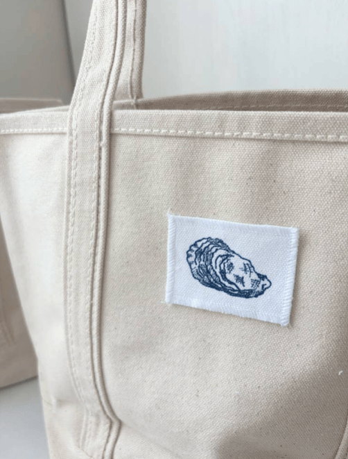 Close-up of a beige canvas tote bag with a white rectangular patch featuring a blue oyster shell illustration.