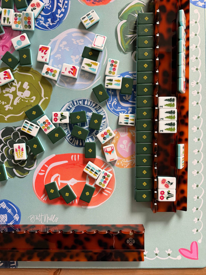 A close-up overhead view of a mahjong game setup with multiple tiles scattered and two aligned stacks on a decorative mat. The mat has a pale blue-green base with colorful printed motifs, and the aligned stacks appear in dark green and a reddish-brown tortoiseshell pattern.