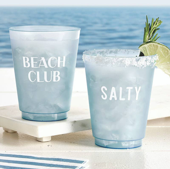 Two blue plastic cups with 'Beach Club' and 'Salty' text, one with a cocktail garnish, on a beach setting.
