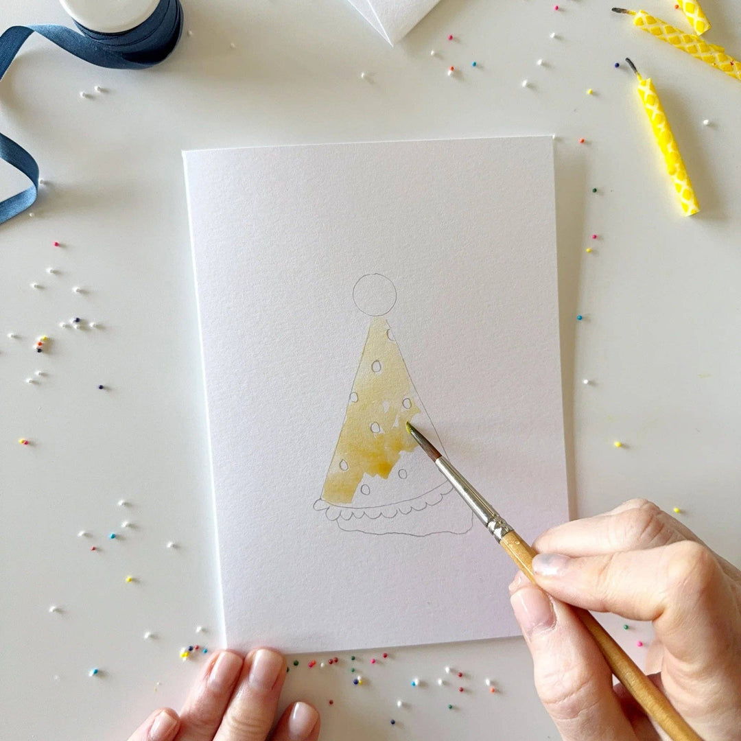 Hand painting a yellow cake with candles on a white card, surrounded by colorful sprinkles and candles.