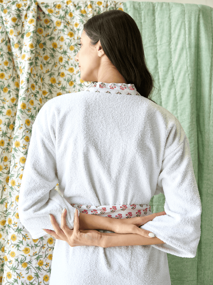 Woman wearing a white robe with floral trim standing in front of a floral curtain and green wall.