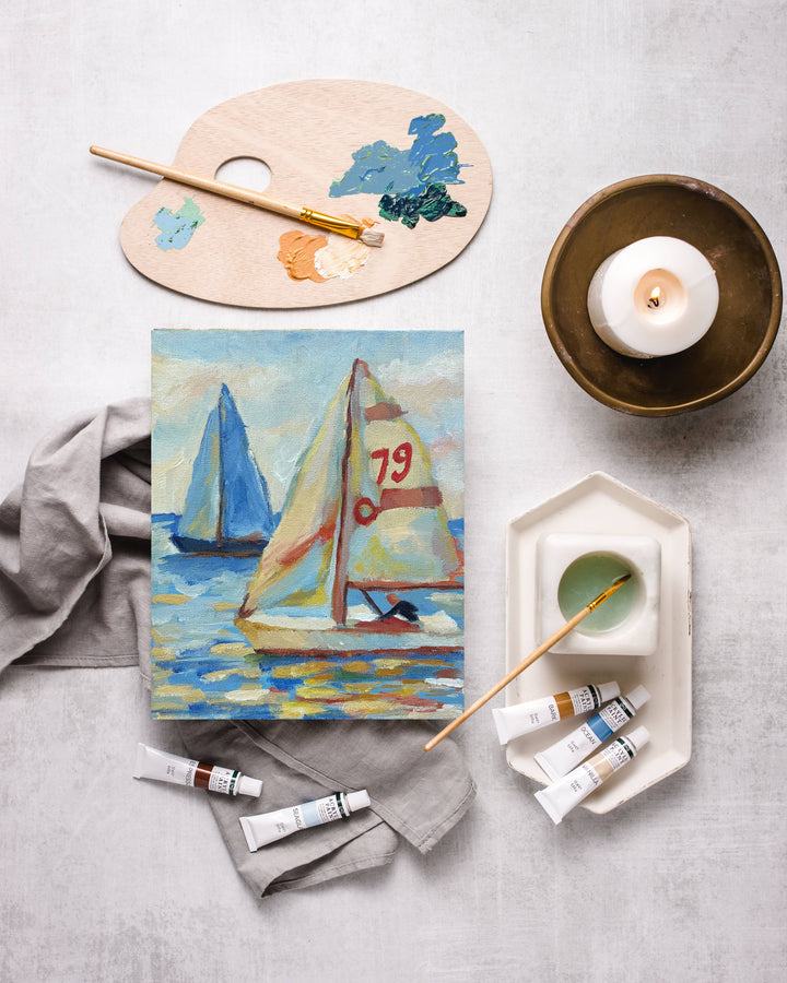 Painting of sailboats on a canvas with art supplies on a gray surface