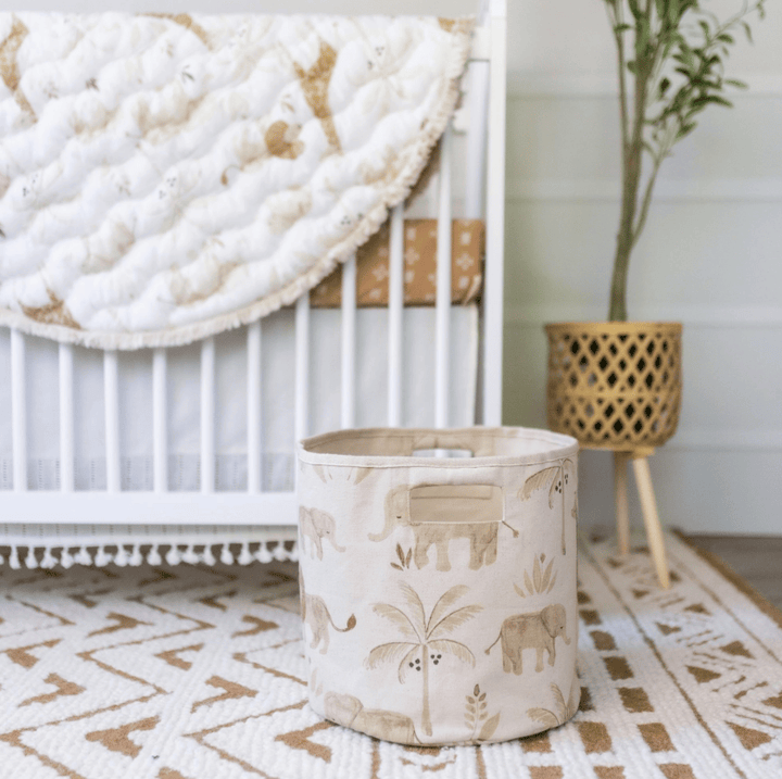 A fabric round storage bin with a beige and white elephant and palm tree pattern, set on a patterned rug in a nursery next to a crib and a potted plant.