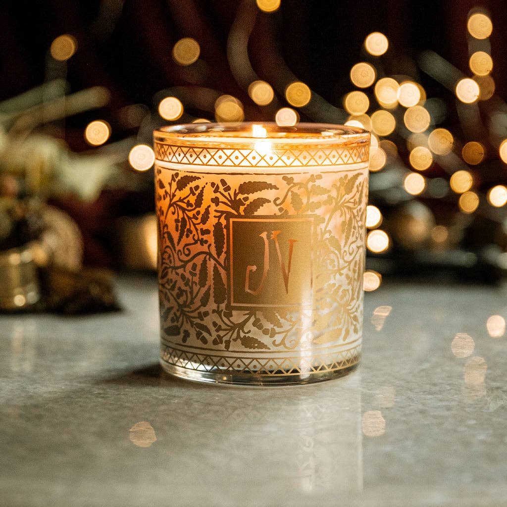 Decorative candle with intricate patterns and initials 'JV' on a reflective surface with blurred lights in the background.