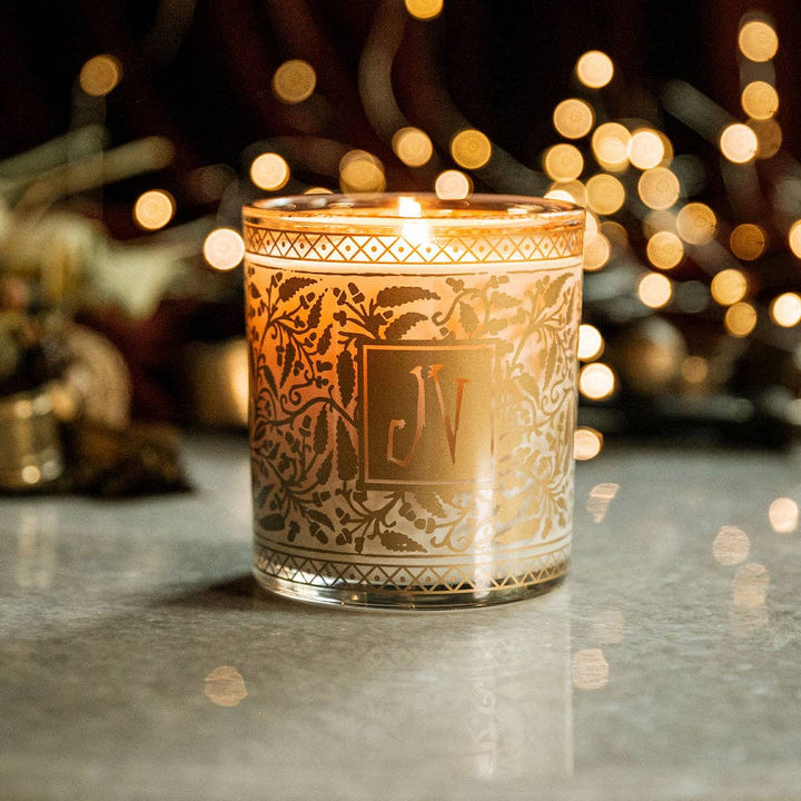 Decorative candle with intricate patterns and initials 'JV' on a reflective surface with blurred lights in the background.
