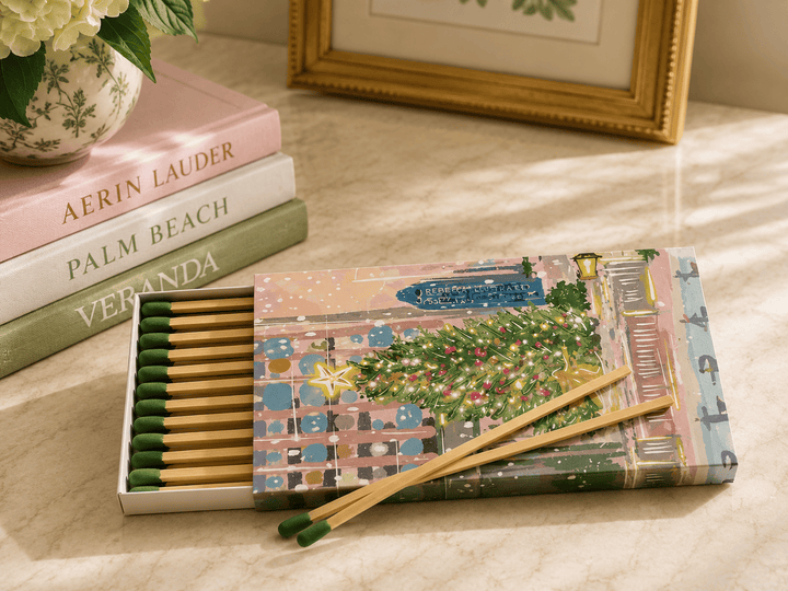 A decorative matchbox set with wooden matches and a matching illustrated cover sits on a light stone surface beside stacked books and a framed picture. The matchbox lid shows a pastel city street scene with a green Christmas tree, and two wooden matches with green tips rest across the front.
