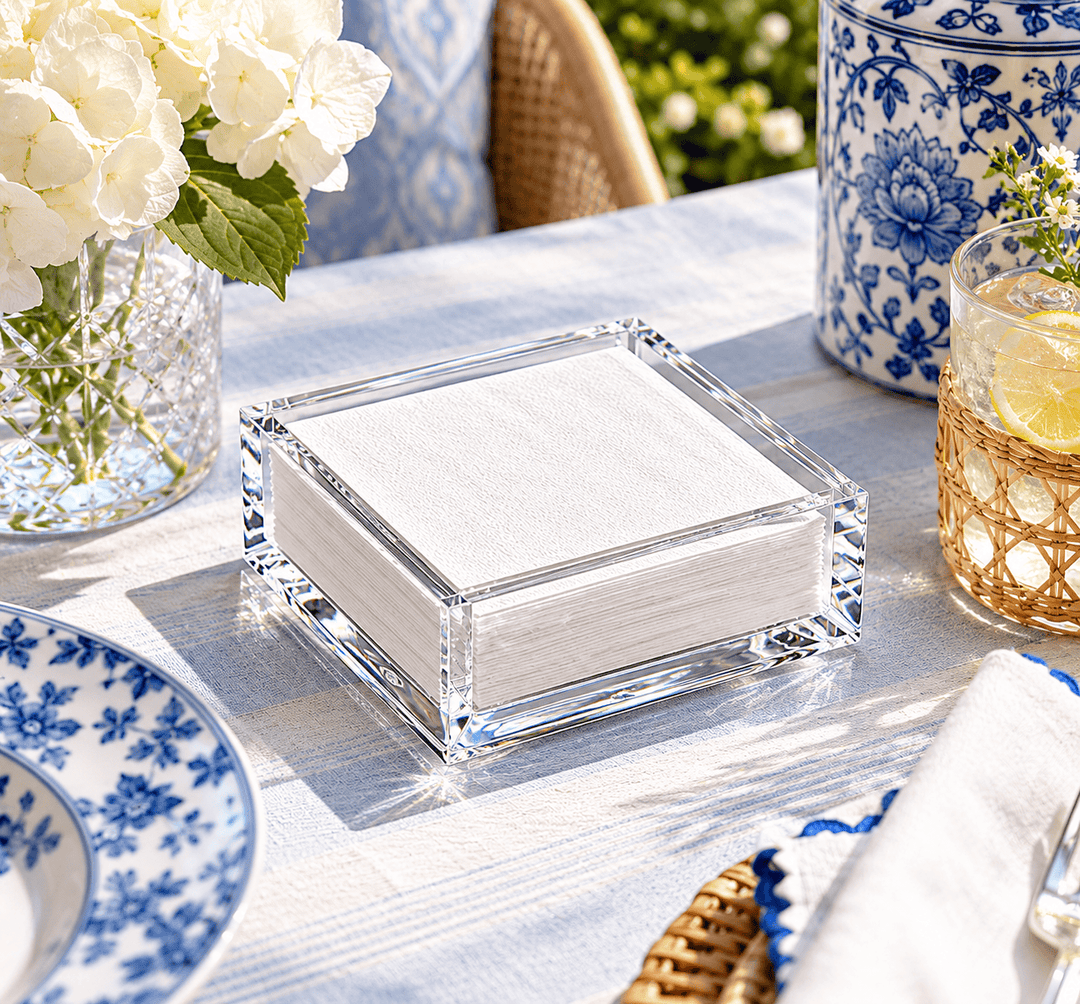 A clear acrylic cocktail napkin holder filled with white napkins, styled on a blue-and-white tablecloth with floral dishes, a vase of white flowers, and a drink with a lemon slice in an outdoor patio setting.