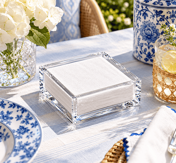 A clear acrylic cocktail napkin holder filled with white napkins, styled on a blue-and-white tablecloth with floral dishes, a vase of white flowers, and a drink with a lemon slice in an outdoor patio setting.
