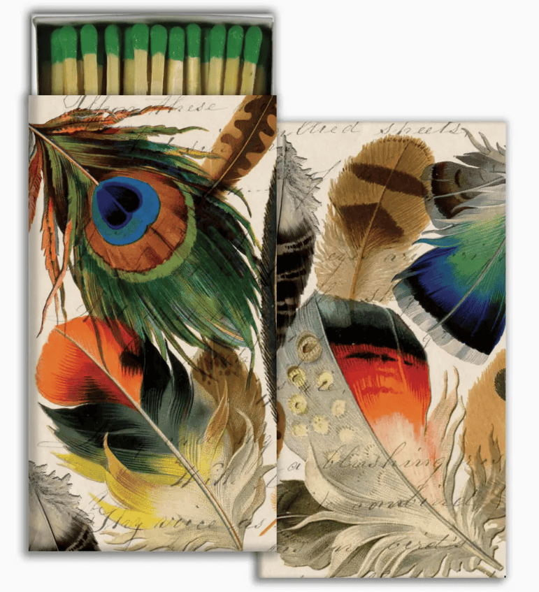 A matchbox-style decorative package with illustrated feathers on the front and a row of green-tipped matches visible in the open top. The design features multicolored feathers including blue, green, orange, yellow, brown, and black on a light cream background with faint script text.