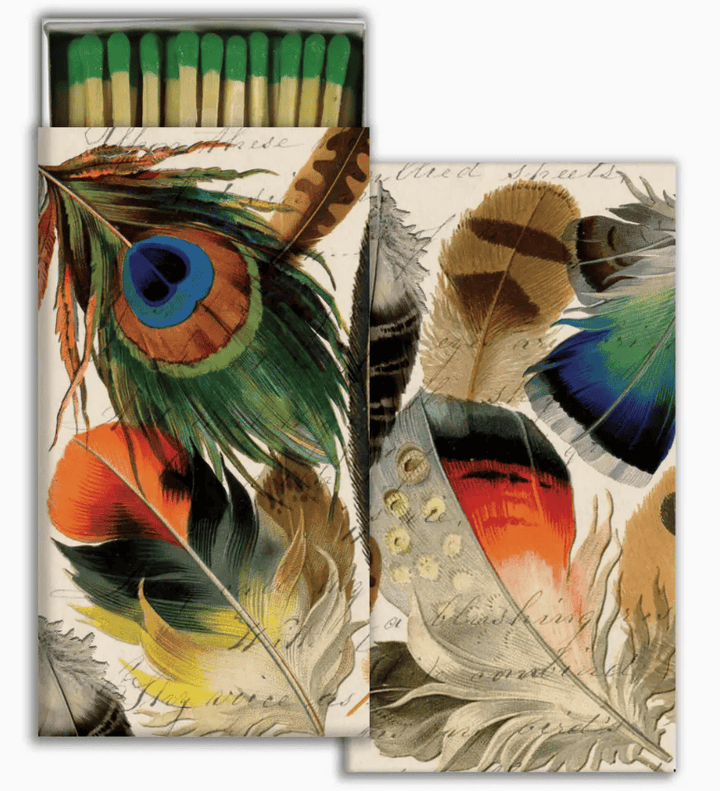 A matchbox-style decorative package with illustrated feathers on the front and a row of green-tipped matches visible in the open top. The design features multicolored feathers including blue, green, orange, yellow, brown, and black on a light cream background with faint script text.