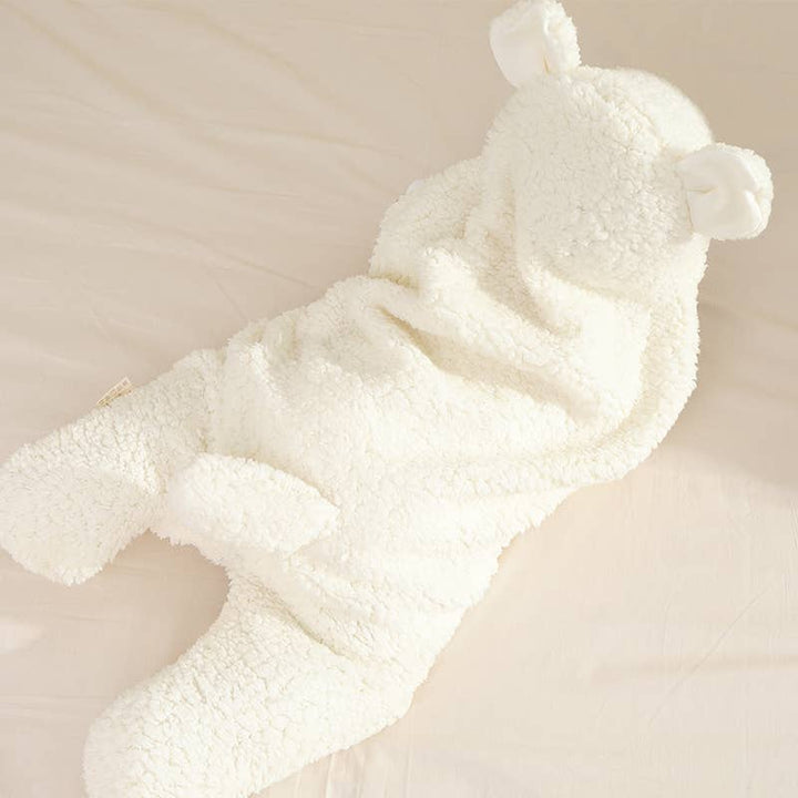 A plush teddy bear-shaped baby onesie in soft cream fleece, laid flat on a light beige fabric background.