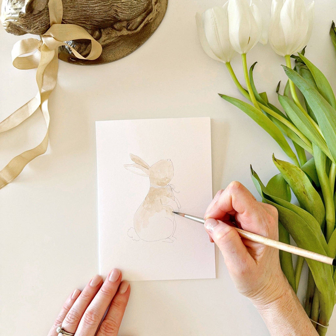Person painting a white tulip on a card with white tulips and ribbons in the background