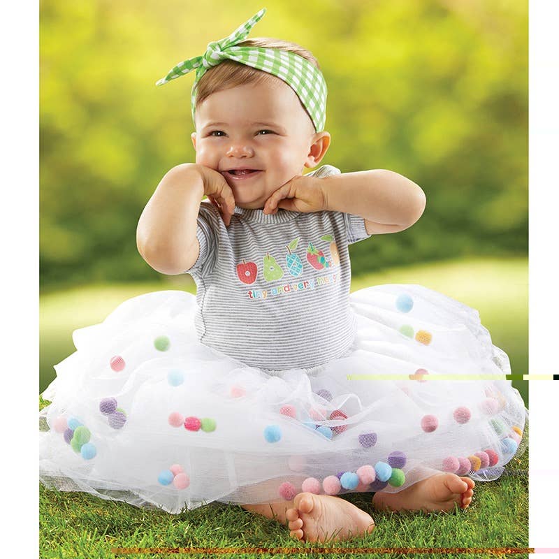 Baby in a striped outfit with a white tutu and colorful pom-poms outdoors.