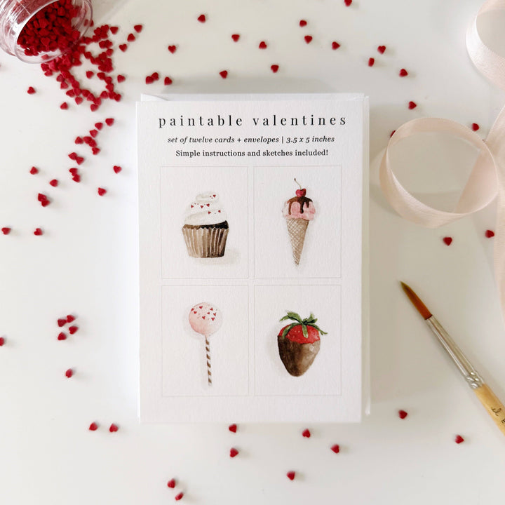 Set of twelve paintable Valentine's cards with illustrations of a cupcake, ice cream cone, balloon, and strawberry on a white card with red heart decorations.