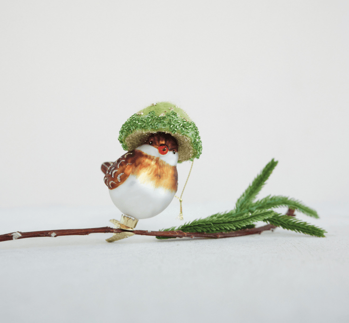 A small bird ornament perched on a branch, wearing a glittery green hat, against a plain light background.
