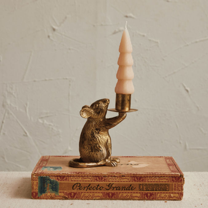 A decorative mouse figurine in metallic gold holding a tiered cream candle, displayed on top of a vintage book against a textured off-white wall.