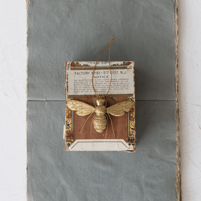 A vintage-style decorative bee ornament mounted on a small printed card, suspended by a thin string against a gray rectangular panel with white walls on either side.