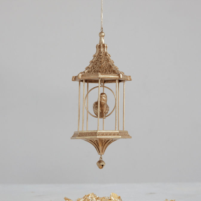 A gold-toned metal bird cage hanging ornament with a small bird figure inside, suspended against a plain light gray background.
