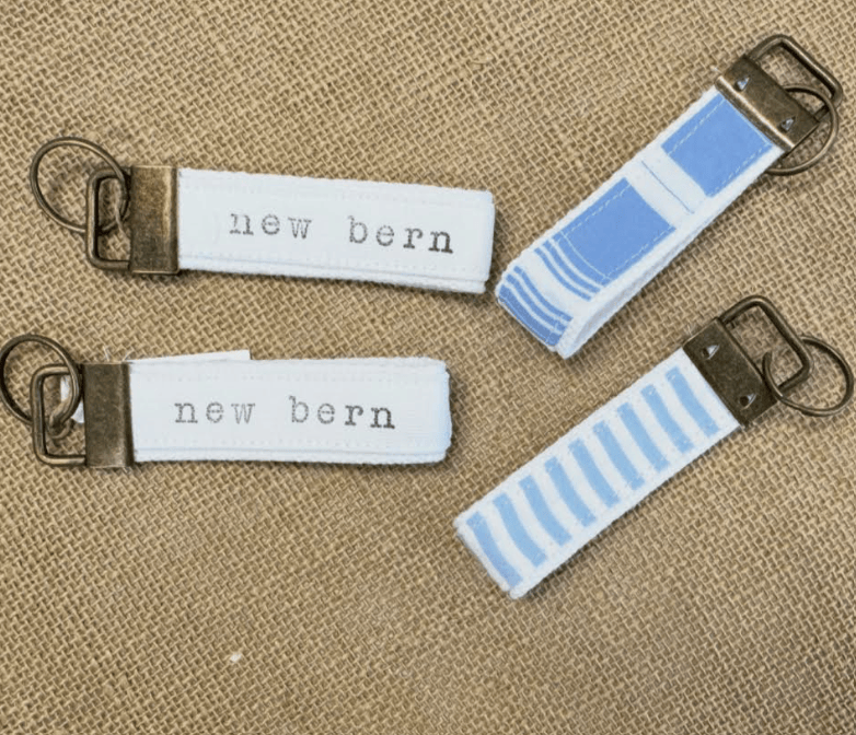 Two fabric keychains displayed on a textured tan burlap background. One pouch is solid white with 'new bern' printed in black, and the other features a light blue and white striped pattern.