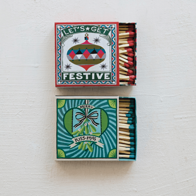 Two decorative matchboxes displayed on a white textured background, one open with red matchheads visible and one closed. The top box reads “LET’S GET FESTIVE” with a colorful ornament illustration, and the bottom box reads “MERRY KISSES-IMAS” with a bow and ribbon illustration.