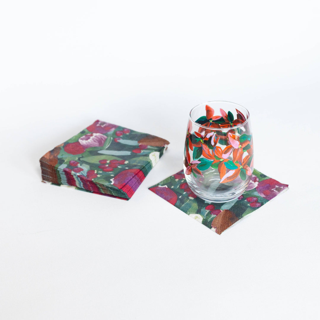 A stack of square paper napkins with a colorful floral and botanical print, shown next to a clear stemless wine glass resting on one napkin. The napkin pattern features red, green, orange, and brown tones on a white background.
