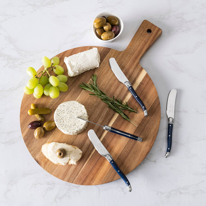 Wooden cutting board with cheese, grapes, olives, and navy blue handled speaders on a light gray surface