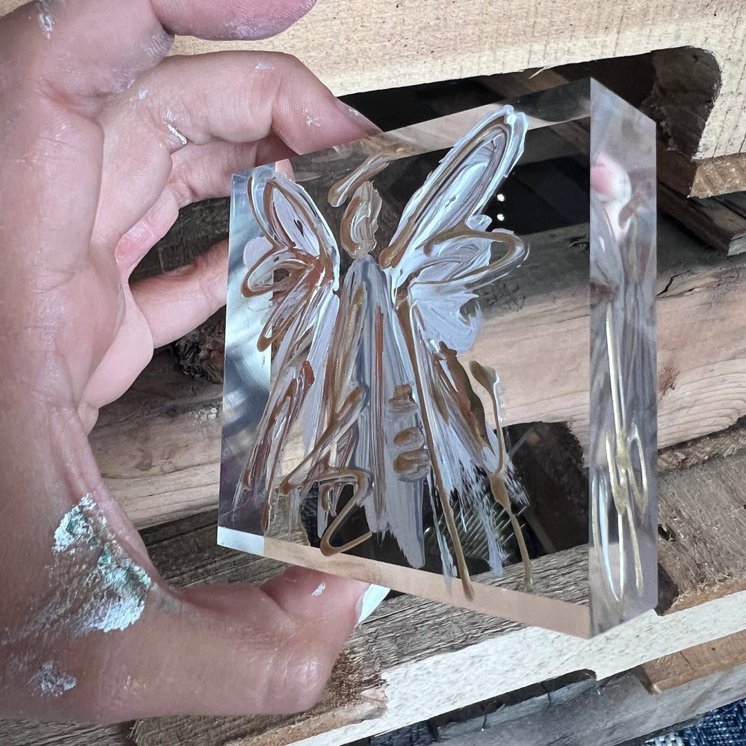 Hand holding a clear acrylic block with an angel design on a wooden surface.