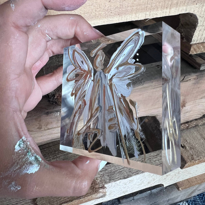 Hand holding a clear acrylic block with an angel design on a wooden surface.